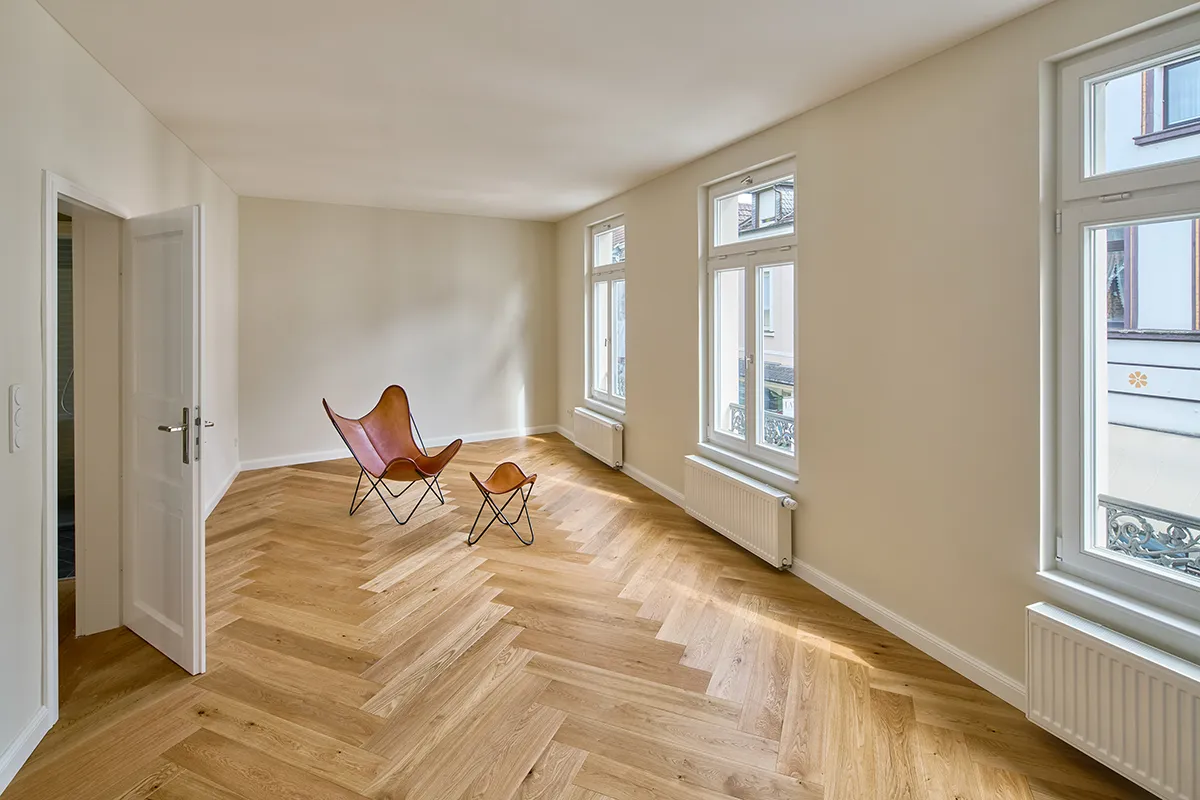 Sanierter Wohnraum mit Fischgrätparkett, hellen Wandflächen und großen Fenstern entlang der Außenfassade.